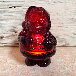 May include: A translucent ruby red glass figurine shaped like Santa Claus. The figure features moulded details including a hat, a thick beard, a textured coat with a belt, and boots. The glass has a deep crimson hue that catches light, creating a bright glow in the centre of the torso area.