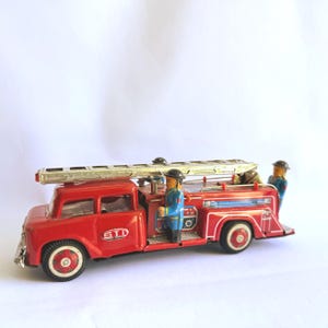Peut inclure: Un jouet vintage en métal rouge représentant un camion de pompiers avec une échelle argentée sur le dessus, des roues blanches à centre rouge et deux petites figurines en uniforme bleu. Le côté affiche les lettres STI dans un ovale blanc.