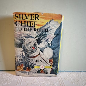 Puede incluir: Portada de libro vintage de "Silver Chief to the Rescue" de Jack O'Brien. La portada presenta un perro blanco con un collar rojo en una escena de montaña nevada. El título está en letras grandes y en negrita.