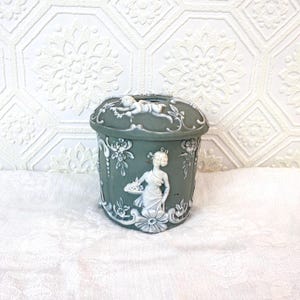 Pode incluir: Um recipiente decorativo verde-azulado com figuras em relevo brancas e desenhos florais. O recipiente apresenta uma tampa com um querubim e uma mulher segurando uma bandeja. O recipiente está sobre uma superfície branca.