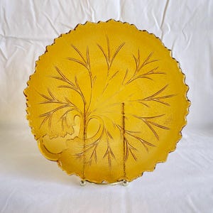 Può includere: Un piatto vintage in vetro color ambra con un design a foglie e rami. Il piatto ha una superficie strutturata e un bordo smerlato. Il disegno della foglia è inciso in un colore ambra più scuro. Il piatto è esposto su un supporto in metallo dorato.