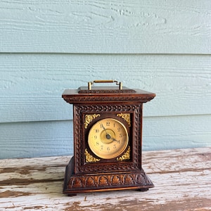 May include: A dark brown wooden mantel clock featuring a square frame with intricate carved detailing, a gold-toned circular clock face with black numerals, and decorative gold-colored metal corner accents. A small metal handle sits on the top.