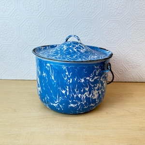 May include: A blue and white enamelware pot with a lid. The pot has a marbled pattern and a black handle. The lid has a small loop handle. The pot is sitting on a wooden surface.