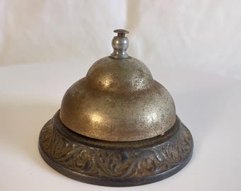 Könnte beinhalten: Antike Messingglocke mit dekorativem Sockel. Die Glocke hat ein angelaufenes, gealtertes Aussehen, mit einem abgerundeten Oberteil und einem kleinen Griff. Der Sockel weist ein verziertes, florales Design auf. Ein klassisches Stück für die Inneneinrichtung.