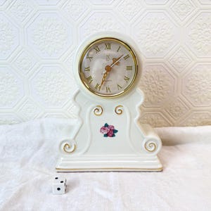 May include: A cream-colored quartz clock with gold Roman numerals and hands. The clock has a decorative base with a floral design and gold accents. A small white die with black dots sits in front of the clock.