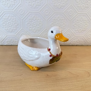 May include: A white ceramic duck-shaped planter with a yellow beak and feet. The planter has a decorative wreath around its neck with red berries and green leaves. The interior of the planter is white.