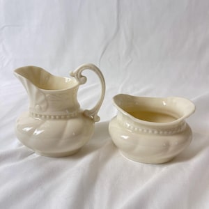 以下が含まれることがあります： クリーム色のセラミック製クリーマーと砂糖入れのセット。クリーマーは湾曲したハンドルと注ぎ口があり、砂糖入れは広い開口部があります。どちらの作品もビーズトリムとキルティング模様が特徴です。