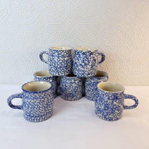 Op de afbeelding: Een verzameling van zeven keramische mokken met een blauw en wit gespikkeld patroon. Elke mok heeft een oor en een licht onregelmatige rand. De mokken zijn gestapeld op een wit oppervlak, met een getextureerde witte achtergrond.