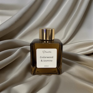 May include: An amber glass bottle with a gold cap. The label reads "Svetlo Cedarwood & Jasmine 100ml". The bottle is square-shaped and sits on a beige, silky fabric.