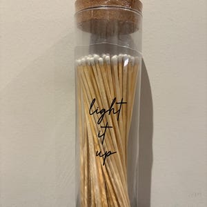 May include: A clear glass jar filled with wooden matches, topped with a cork lid. The jar has the words "light it up" in a handwritten font. The matches have white tips.
