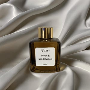 Może przedstawiać: Kwadratowy, bursztynowy flakon perfum ze złotą nakrętką. Etykieta głosi "Svetlo Musk & Sandalwood 100ml". Butelka stoi na białej, jedwabistej tkaninie.