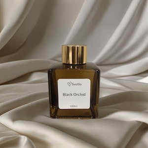 May include: A brown glass perfume bottle with a gold cap. The label reads "Svetlo Black Orchid 100ml". The bottle is square and sits on a cream-coloured, draped fabric background.