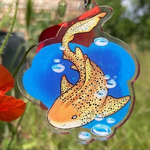 Peut inclure: Un porte-clés en acrylique transparent avec un motif de requin léopard. Le requin est orange et jaune avec des taches noires, sur un fond bleu avec des bulles. Le porte-clés a un accent rouge en haut et un anneau argenté.