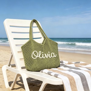 Puede incluir: Bolso tote de crochet verde oliva con el nombre "Olivia" en escritura blanca. La bolsa est&aacute; sobre una silla de playa blanca, con una toalla a rayas. El fondo muestra una playa de arena y el mar.