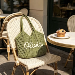 Puede incluir: Un bolso tote de ganchillo verde con el nombre "Olivia" en escritura blanca se muestra en una silla de mimbre. El bolso tiene una abertura ancha y asas largas. Un muffin con ar&aacute;ndanos est&aacute; en un plato peque&ntilde;o sobre una mesa.