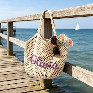 Puede incluir: Un bolso tote de crochet beige con el nombre "Olivia" bordado en morado. El bolso cuelga de una barandilla de madera, con gafas de sol y flores rosas en su interior. El fondo muestra un muelle, el oc&eacute;ano y un velero.