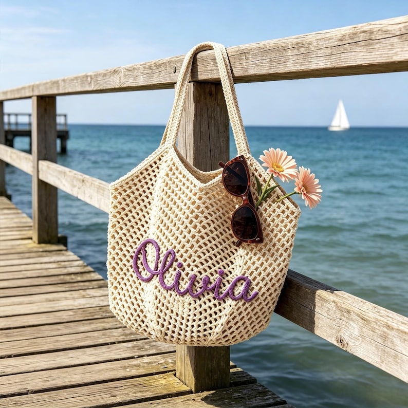Puede incluir: Un bolso tote de crochet beige con el nombre "Olivia" bordado en morado. El bolso cuelga de una barandilla de madera, con gafas de sol y flores rosas en su interior. El fondo muestra un muelle, el oc&eacute;ano y un velero.