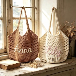 Puede incluir: Dos bolsas de mano de crochet colgadas cerca de una ventana. Una es de color marr&oacute;n con "Anna" bordado en blanco, la otra es blanquecina con "Lily" en rosa. Ambas tienen asas largas y un tejido similar a una red.
