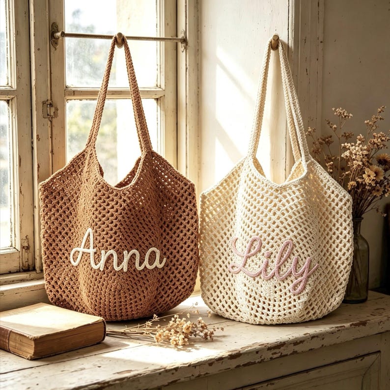 Puede incluir: Dos bolsas de mano de crochet colgadas cerca de una ventana. Una es de color marr&oacute;n con "Anna" bordado en blanco, la otra es blanquecina con "Lily" en rosa. Ambas tienen asas largas y un tejido similar a una red.