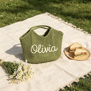 Puede incluir: Un bolso tote verde tejido a ganchillo con el nombre "Olivia" en escritura blanca. El bolso est&aacute; sobre una manta blanca, junto a un ramo de margaritas y un plato con s&aacute;ndwiches. La escena est&aacute; ambientada al aire libre.