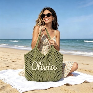 Puede incluir: Bolso tote de crochet verde oliva con el nombre "Olivia" en escritura blanca. El bolso tiene un dise&ntilde;o de tejido abierto grande y asas largas. El bolso est&aacute; sobre una toalla blanca en una playa de arena.