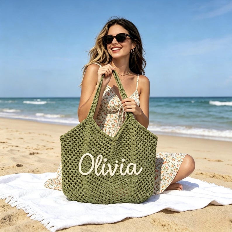 Puede incluir: Bolso tote de crochet verde oliva con el nombre "Olivia" en escritura blanca. El bolso tiene un dise&ntilde;o de tejido abierto grande y asas largas. El bolso est&aacute; sobre una toalla blanca en una playa de arena.