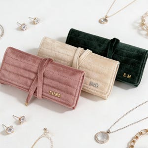Peut inclure: Trois trousses à bijoux en velours rose poudré, crème et vert foncé. Chaque trousse est fermée par un lien assorti et comporte des lettres dorées avec des noms ou des initiales. Des bijoux, dont des boucles d'oreilles et des colliers, entourent les trousses.