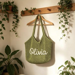 Puede incluir: Un bolso tote de ganchillo verde con el nombre "Olivia" en escritura blanca. El bolso est&aacute; colgado de una percha de madera contra una pared blanca, con plantas en el fondo.