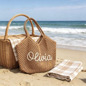 Puede incluir: Un bolso de playa tejido marr&oacute;n con el nombre "Olivia" en escritura blanca, una cesta de picnic de mimbre y una manta a cuadros doblada en una playa de arena. El oc&eacute;ano es visible en el fondo.