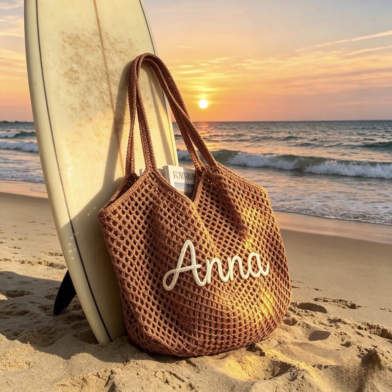 Puede incluir: Un bolso tote marr&oacute;n de tejido abierto con el nombre "Anna" en escritura blanca. El bolso est&aacute; apoyado contra una tabla de surf en una playa de arena al atardecer. El oc&eacute;ano y el cielo son visibles en el fondo.
