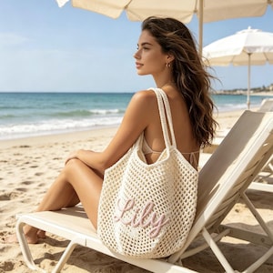 Puede incluir: Bolso tote de tejido abierto color crema con el nombre "Lily" bordado en rosa. El bolso tiene dos correas para los hombros y se muestra en una playa, lo que sugiere un tema de verano o vacaciones. El bolso es lo suficientemente grande para llevar lo esencial de la playa.