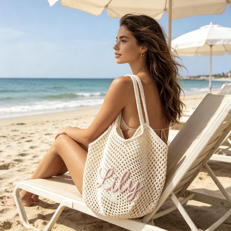Puede incluir: Bolso tote de tejido abierto color crema con el nombre "Lily" bordado en rosa. El bolso tiene dos correas para los hombros y se muestra en una playa, lo que sugiere un tema de verano o vacaciones. El bolso es lo suficientemente grande para llevar lo esencial de la playa.