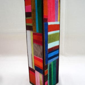 May include: A tall, rectangular decorative vase with a patchwork design. The vase is covered in various colorful fabric patches, including pink, red, green, and blue. The edges are stitched with dark brown thread, creating a unique, handcrafted look.