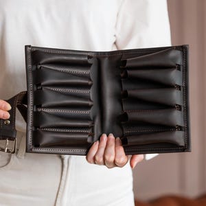 Puede incluir: Estuche enrollable de cuero marrón oscuro para cuchillos con múltiples ranuras individuales. El estuche está abierto, revelando los bolsillos interiores. Se adjunta un cinturón de cuero a juego con una hebilla plateada.