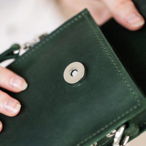 May include: Close-up of a dark green leather crossbody bag with a silver magnetic clasp. The bag's interior is visible, revealing a matching green lining. The stitching is visible around the edges of the bag.