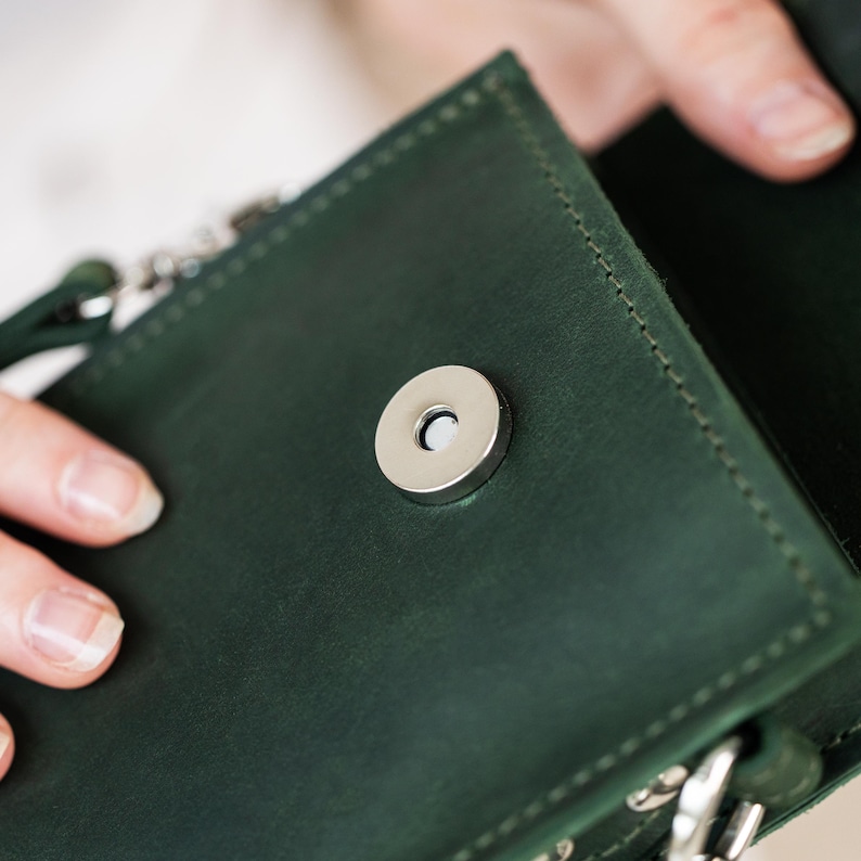 May include: Close-up of a dark green leather crossbody bag with a silver magnetic clasp. The bag's interior is visible, revealing a matching green lining. The stitching is visible around the edges of the bag.