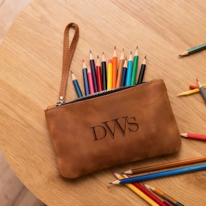 May include: A brown leather pencil case with a zip, wrist strap, and the initials "DWS" engraved on the front. The case is filled with a variety of coloured pencils. Some pencils are scattered on the wooden surface.