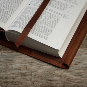 May include: An open book with a brown leather cover and a matching bookmark. The pages are white with black text. The leather has a rich, warm tone, and the bookmark is placed between the pages.