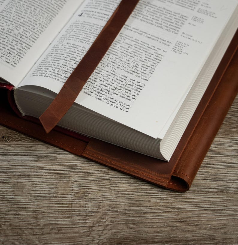 May include: An open book with a brown leather cover and a matching bookmark. The pages are white with black text. The leather has a rich, warm tone, and the bookmark is placed between the pages.