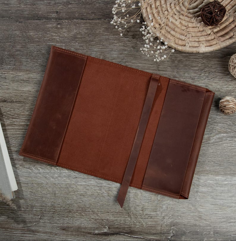 May include: Open, brown leather journal with a matching bookmark. The journal has a textured, distressed appearance. The cover is a darker shade of brown than the interior pages. The journal is lying open on a wooden surface.