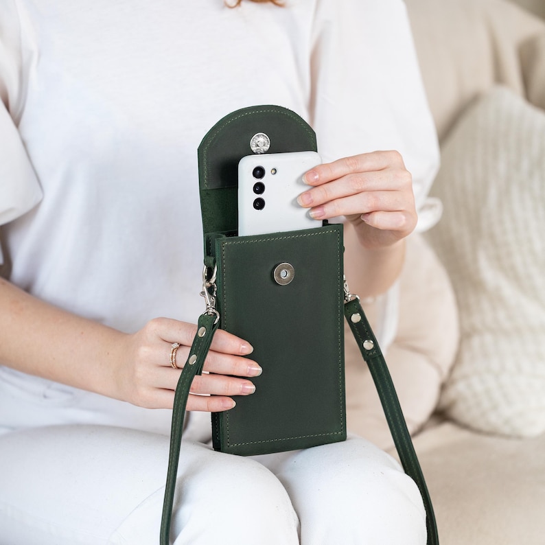 May include: A dark green leather crossbody phone bag with a flap closure and a detachable strap. The bag is designed to hold a smartphone, and a white smartphone is partially visible inside. The bag has a magnetic snap closure and silver-tone hardware.