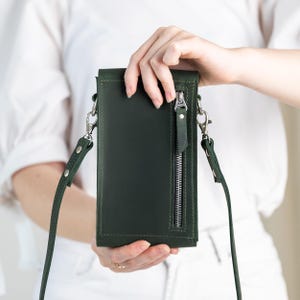 May include: A dark green leather crossbody phone bag with a silver zipper and strap hardware. The bag has a long, adjustable strap and a vertical zipper pocket on the front. The bag is held by a person wearing a white shirt and white pants.