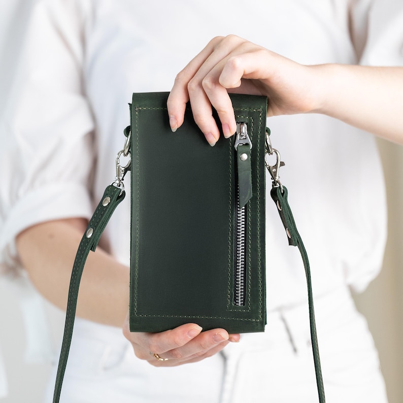 May include: A dark green leather crossbody phone bag with a silver zipper and strap hardware. The bag has a long, adjustable strap and a vertical zipper pocket on the front. The bag is held by a person wearing a white shirt and white pants.