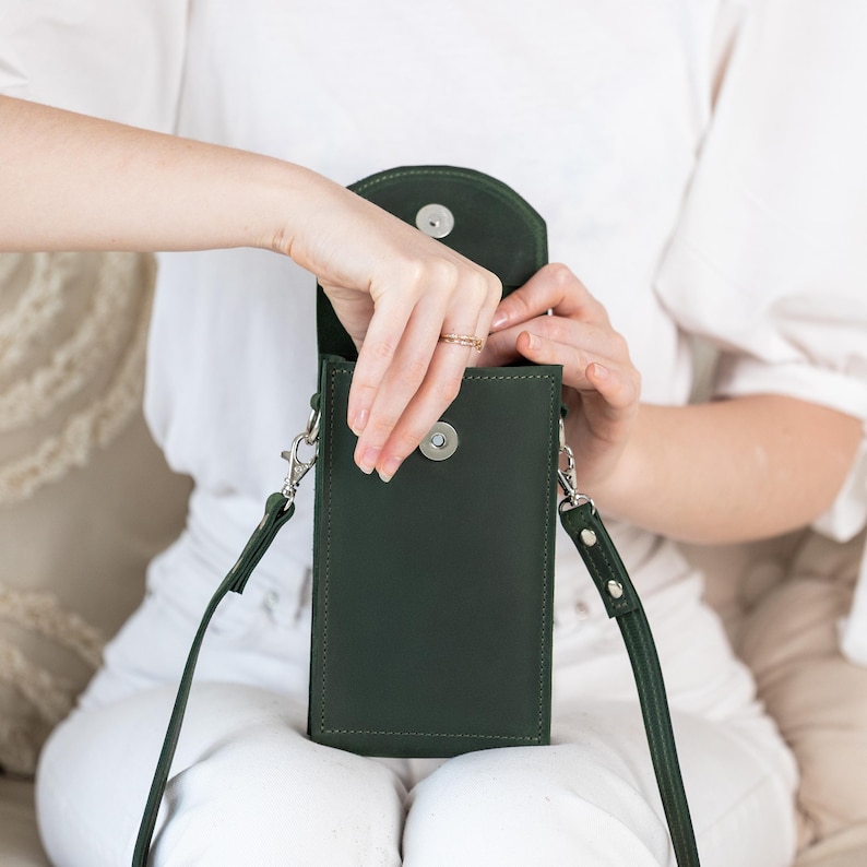 May include: A dark green leather crossbody phone bag with a long strap and silver-tone hardware. The bag has a rectangular shape with a flap closure. The bag is being opened to reveal the interior.