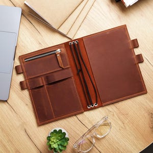 May include: Open brown leather journal with a zip pocket, pen holders, and elastic closure. The journal is on a wooden desk with a laptop, glasses, and a small succulent plant.