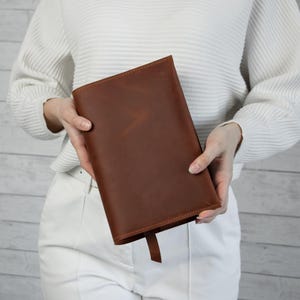 May include: A brown leather book cover with a matching bookmark. The cover is rectangular and appears to be made of high-quality leather. The leather has a natural grain and a warm, reddish-brown color. The bookmark is a simple, thin strip of the same leather.