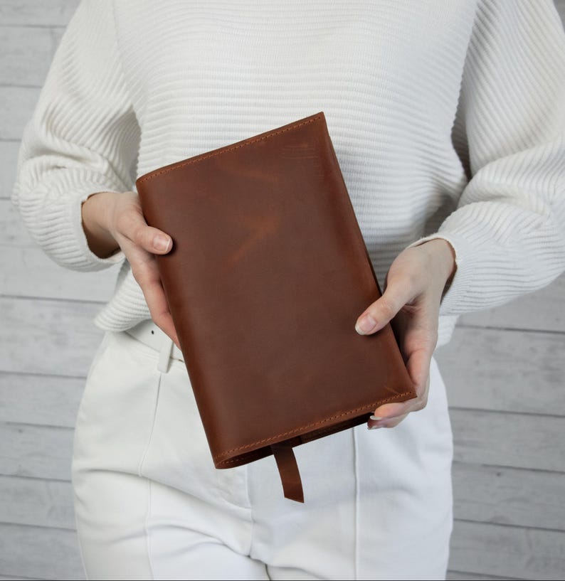 May include: A brown leather book cover with a matching bookmark. The cover is rectangular and appears to be made of high-quality leather. The leather has a natural grain and a warm, reddish-brown color. The bookmark is a simple, thin strip of the same leather.
