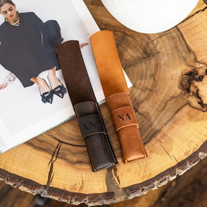 Puede incluir: Dos estuches de cuero para bolígrafos en marrón y beige, cada uno con una correa e iniciales en relieve. El estuche marrón tiene "AN" en relieve, y el estuche beige tiene "NA". Los estuches están sobre una superficie de madera.