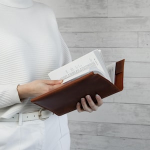 May include: A brown leather book cover holds an open book with white pages. The cover is a rich, warm brown color. The book is held open, revealing the text within. The background is a light wood-paneled wall.