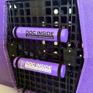 May include: A purple glittery RuffLand Performance Kennels crate featuring a black grid door. Two purple cylindrical containers are attached to the door, each labeled with white text reading DOG INSIDE EMERGENCY INFORMATION TWIST OPEN.